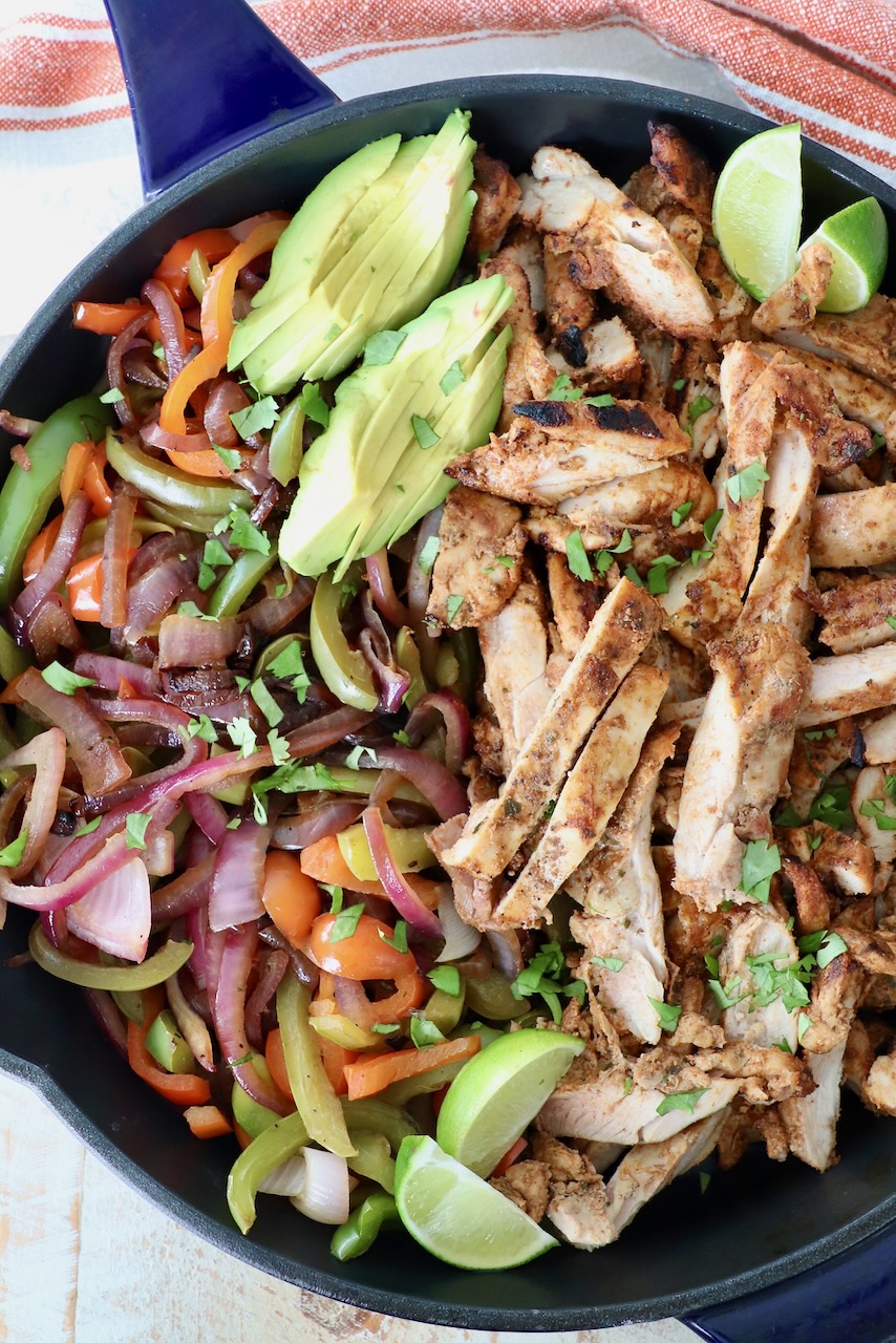 grilled chicken and vegetables in skillet with sliced avocado and lime wedges