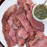 thinly sliced cooked picanha steak on plate with a small bowl of chimichurri sauce on the side