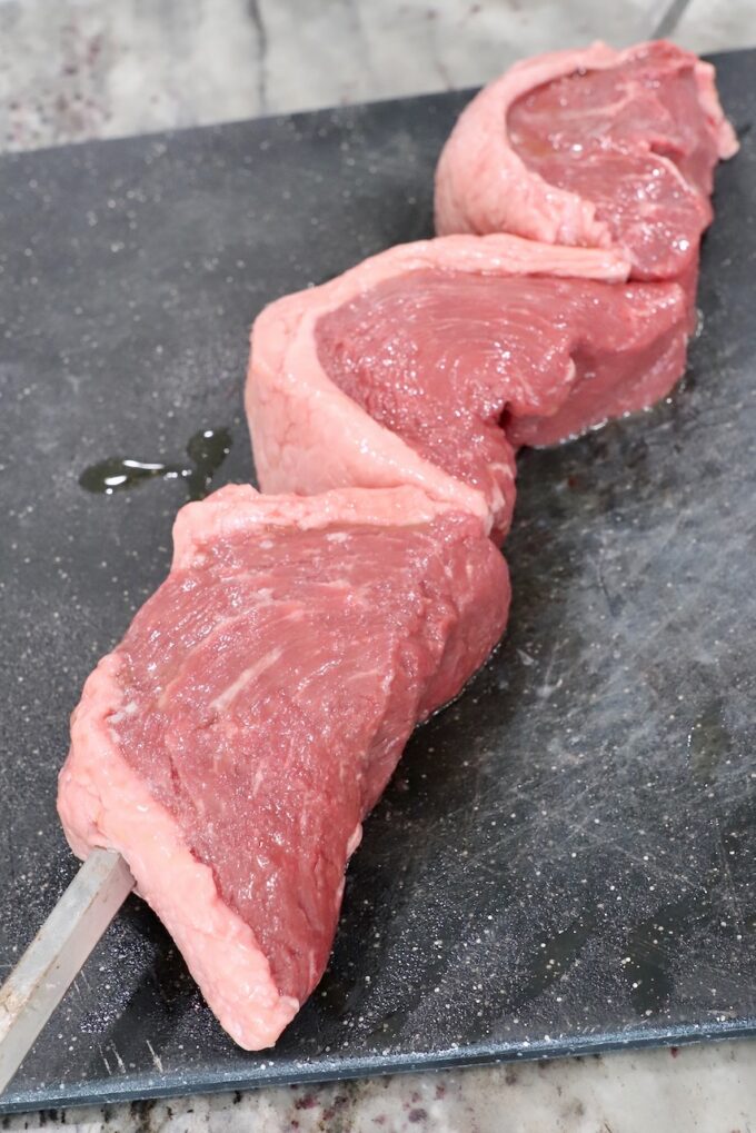 three pieces of steak skewered on a rotisserie rod on a cutting board