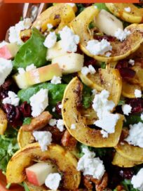 salad in bowl topped with roasted strips of delicata squash, chopped pecans and crumbled goat cheese