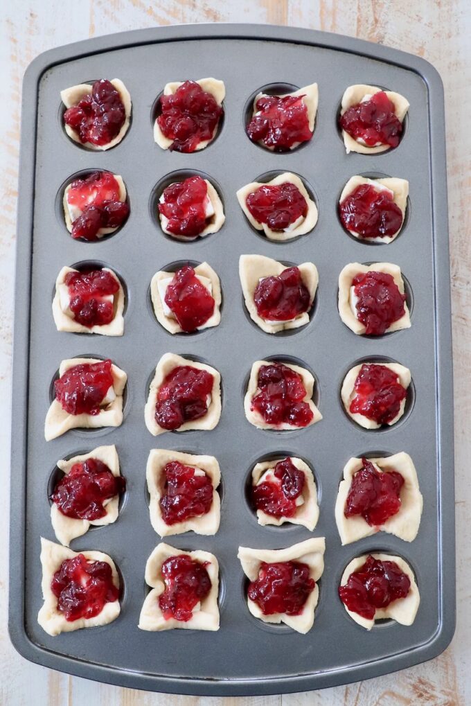 crescent roll dough pressed into a mini muffin tin, topped with pieces of brie cheese and cranberry sauce