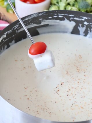 pot of fondue with a cube of salami and a cherry tomato dipped in the cheese