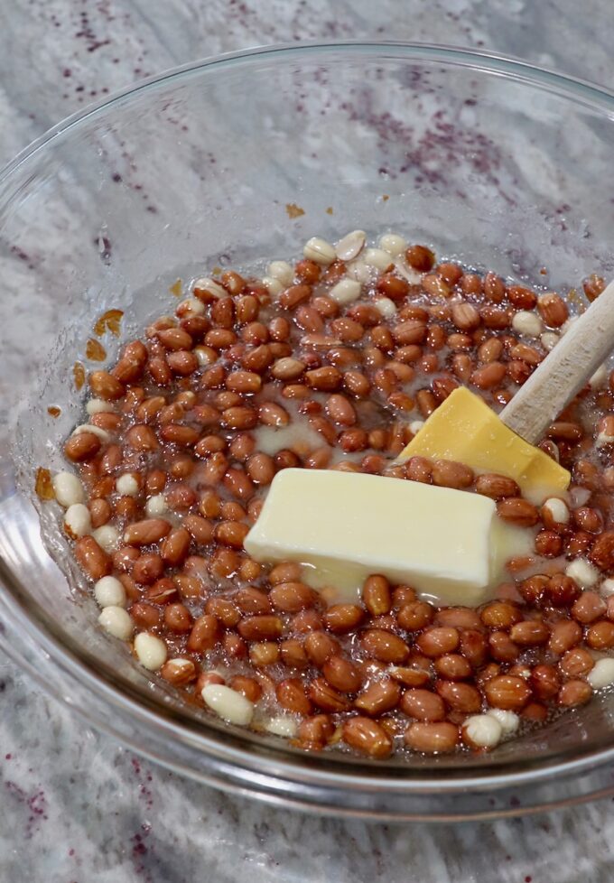 spatula stirring a stick of butter with microwaved sugar, corn syrup and raw peanuts in a large glass bowl