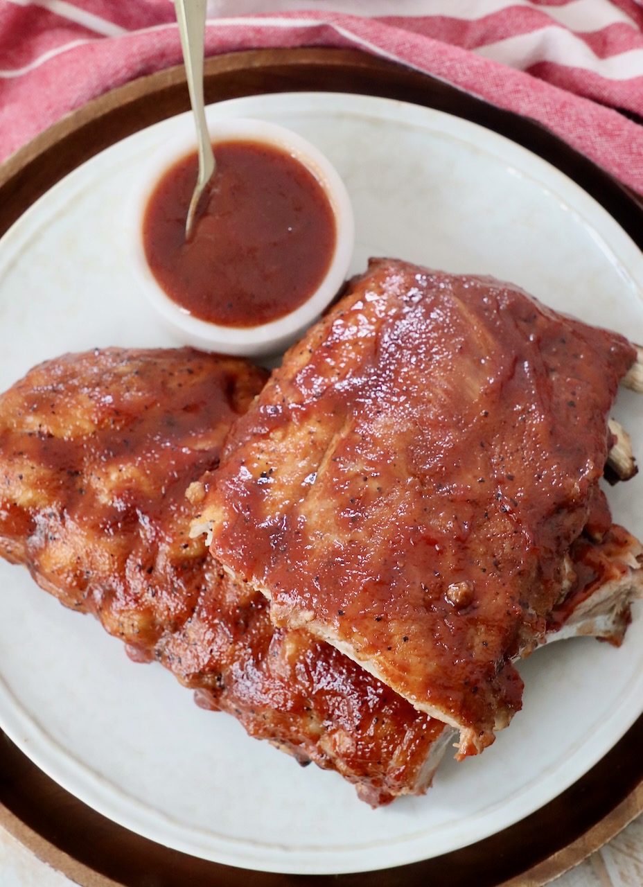 rack of cooked baby back ribs, cut in half on plate with a small bowl of bbq sauce