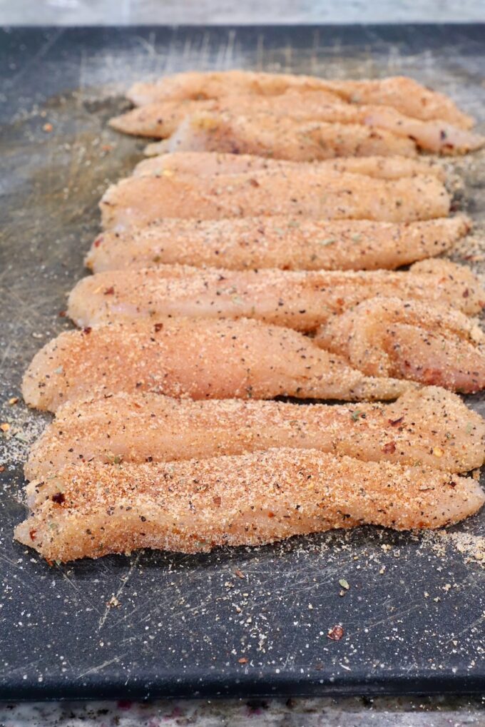 strips of chicken breast meat tossed with seasoning on a cutting board