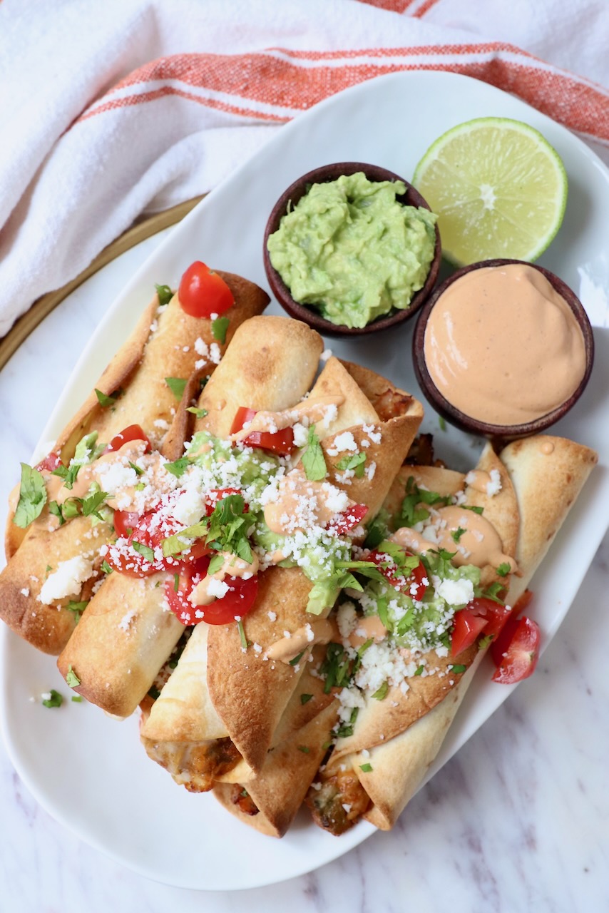 taquitos rolled up on plate topped with cotija cheese, diced tomatoes and guacamole