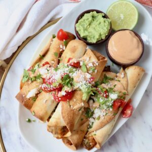 cooked taquitos stacked up on plate with guacamole and chipotle sauce
