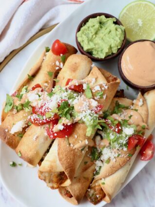 cooked taquitos stacked up on plate with guacamole and chipotle sauce