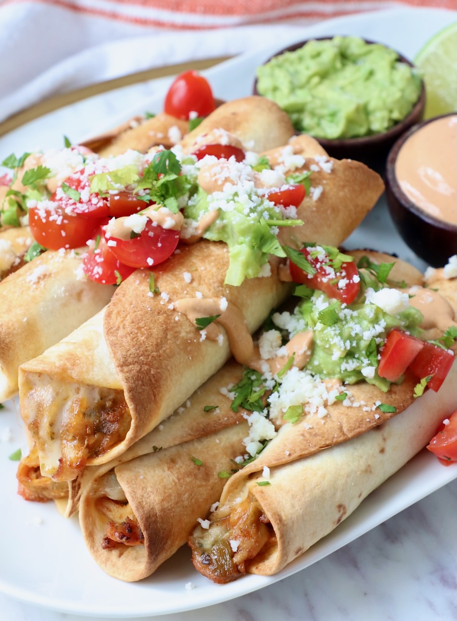 cooked taquitos stacked up on plate, topped with diced tomatoes, cilantro and cotija cheese