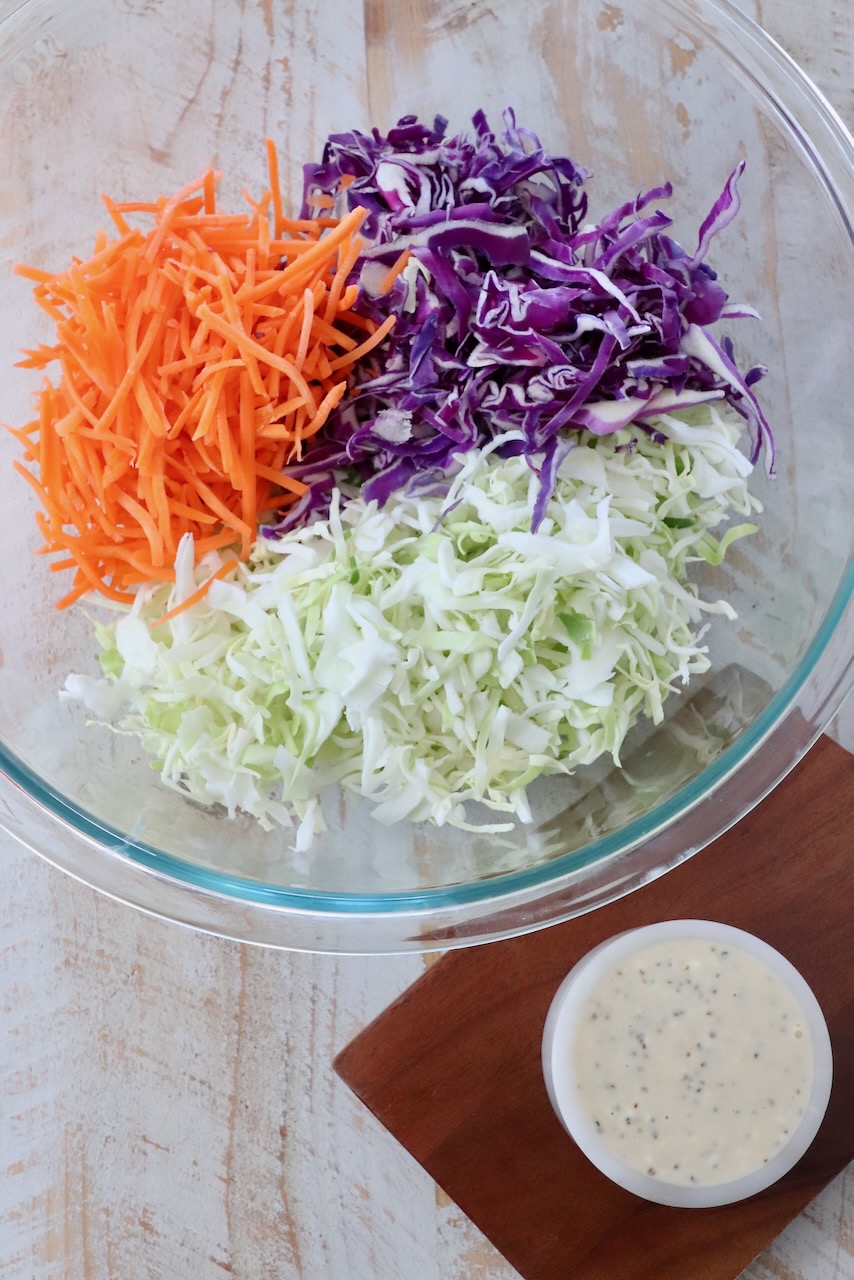 shredded cabbage and carrots in large glass bowl next to a small bowl of coleslaw dressing