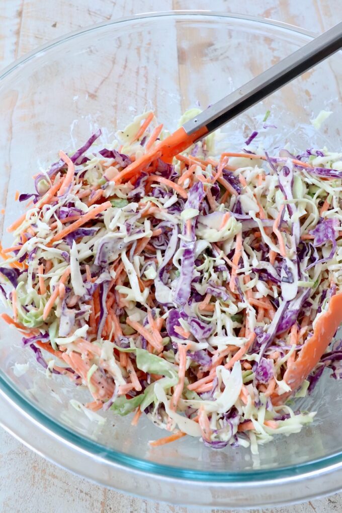 prepared coleslaw in large glass bowl with kitchen tongs