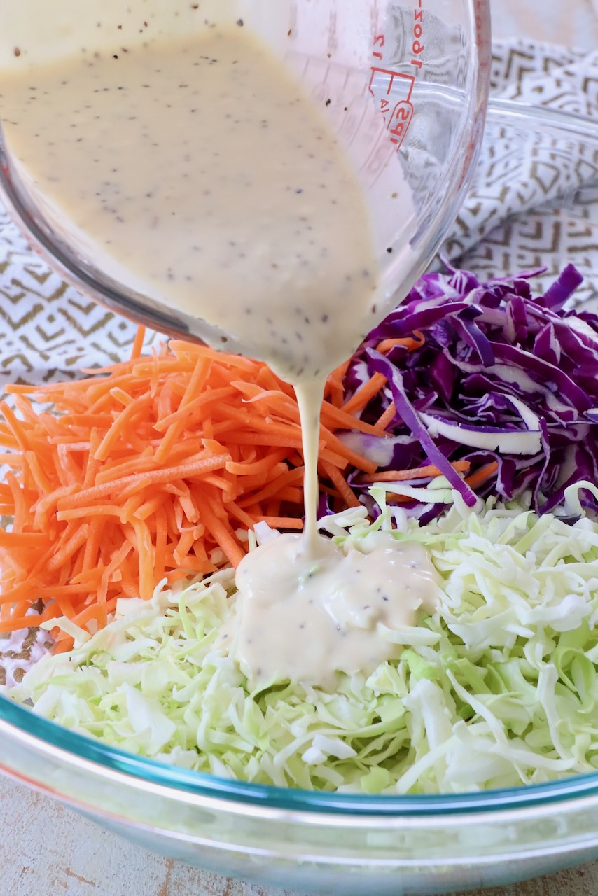 coleslaw dressing poured from glass measuring cup over cabbage in a large glass bowl