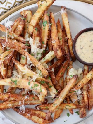 cooked fries on plate topped with parmesan cheese and parsley, with small bowl of sauce on the side