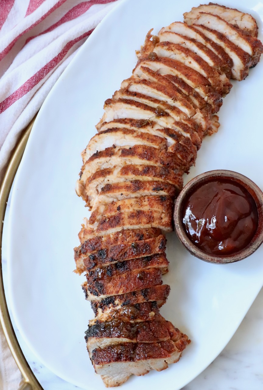 sliced grilled pork tenderloin on plate with a small bowl of bbq sauce on the side