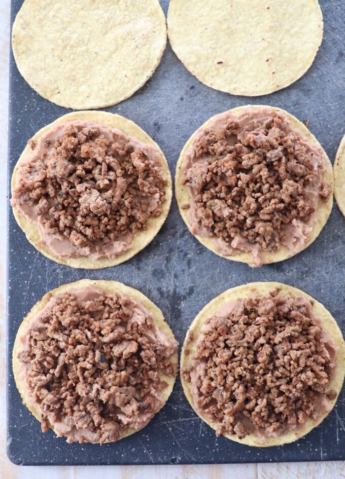 tostada shells topped with refried beans and ground beef on a cutting board