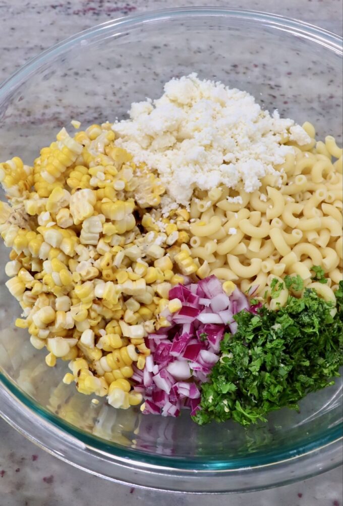 cooked pasta in large glass bowl with diced onions, cilantro, corn kernels and cotija cheese