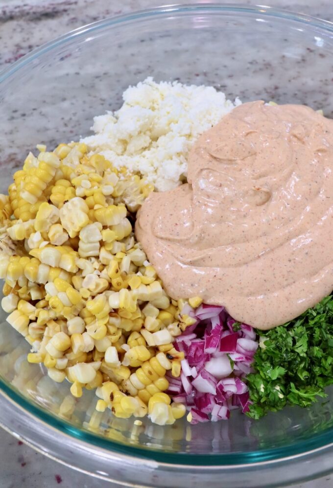 cooked pasta in large glass bowl with diced onions, cilantro, corn kernels, creamy dressing and cotija cheese