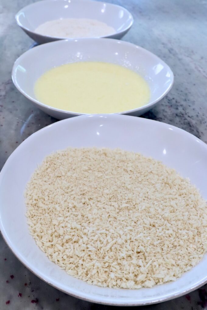 three bowls lined up, one filled with breadcrumbs, one with flour and another with buttermilk