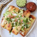 taquitos stacked up on plate topped with guacamole, pico de gallo and cilantro