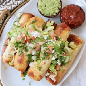 taquitos stacked up on plate topped with guacamole, pico de gallo and cilantro