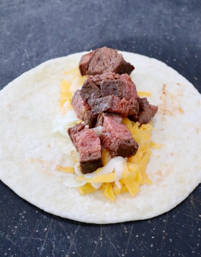 tortilla on cutting board topped with shredded cheese and diced grilled steak