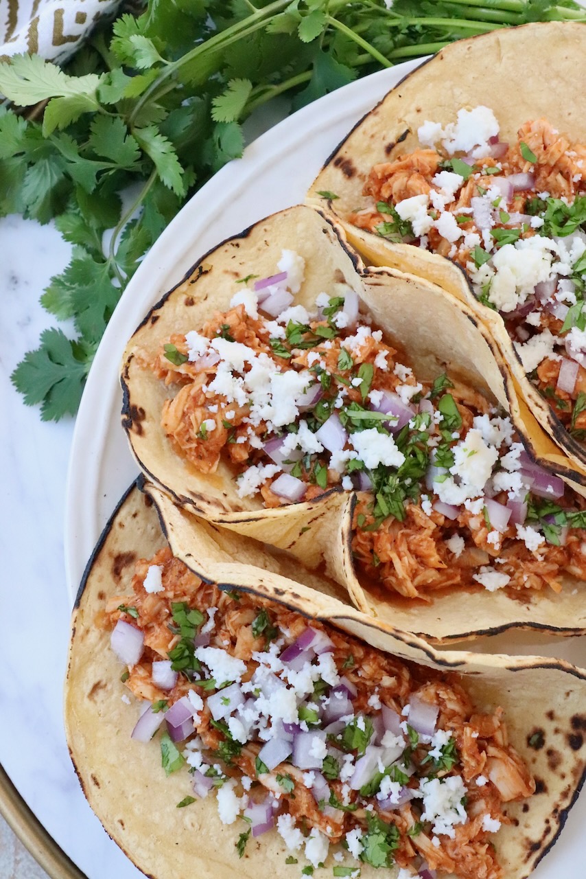 three tacos on plate filled with chicken tinga, cotija cheese, cilantro and onions