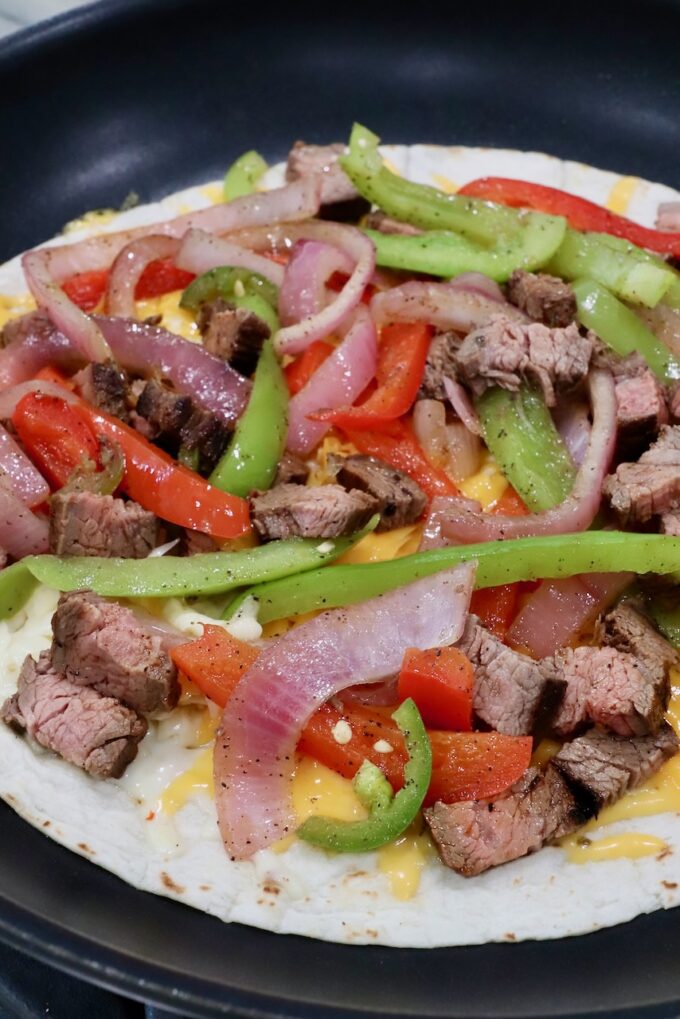 fajita veggies and grilled steak on top of shredded cheese on tortilla in skillet