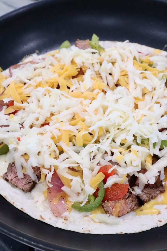 shredded cheese on top of fajita veggies and grilled steak on tortilla in skillet