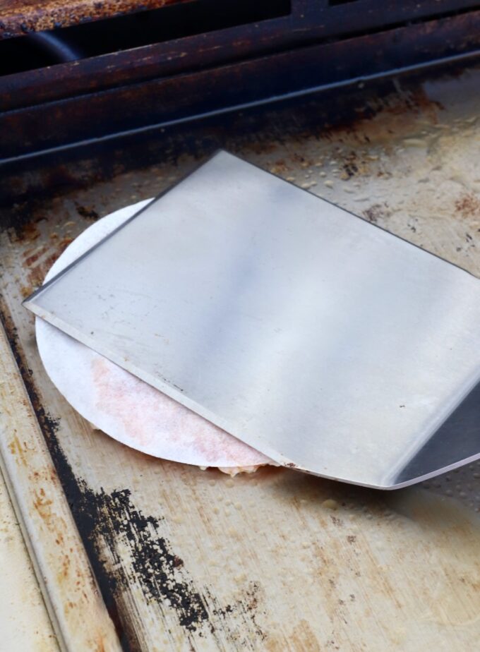 spatula pressing on a chicken burger patty, topped with a piece of parchment paper on a griddle