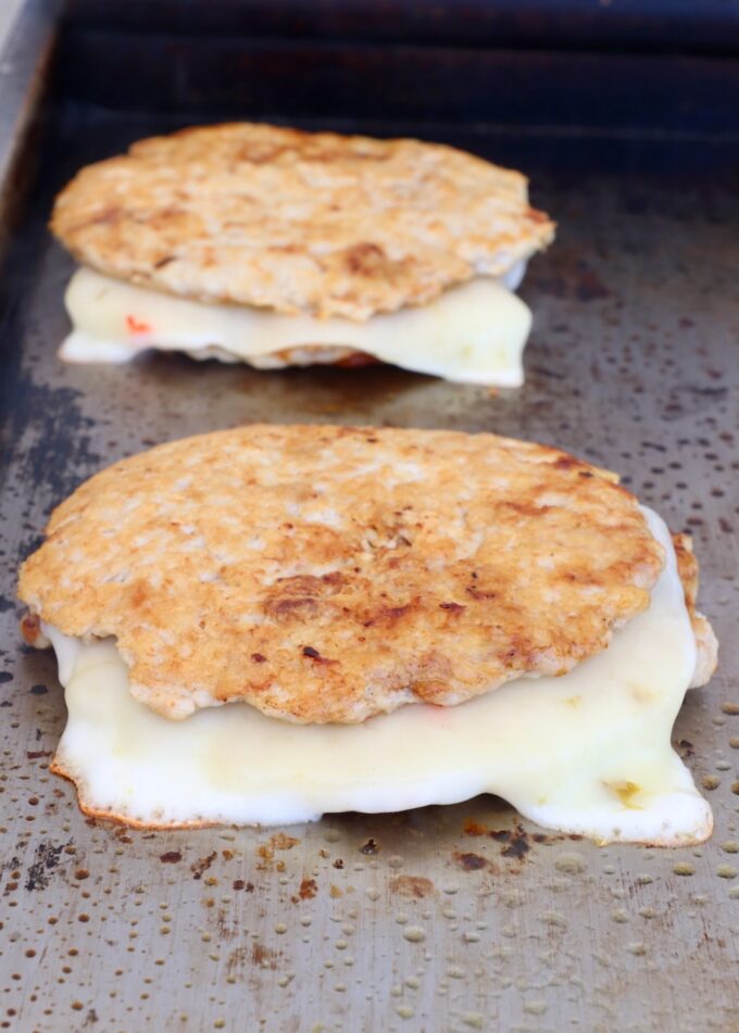 cooked chicken burger patties stacked up with slices of pepper jack cheese melted in the middle on a griddle