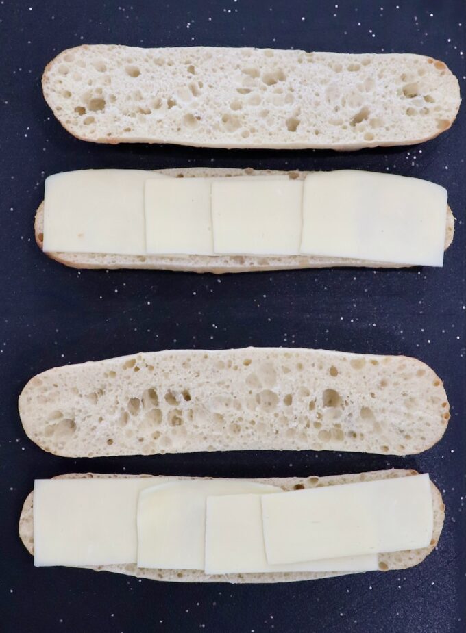 two demi baguettes sliced in half on cutting board, topped with sliced mozzarella cheese