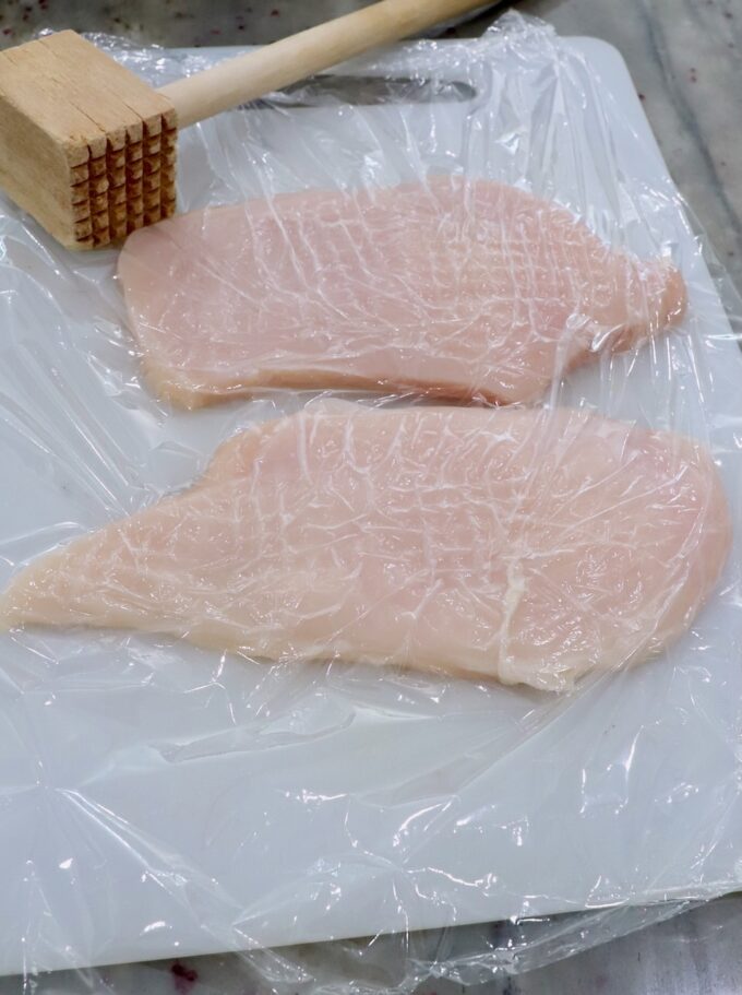 chicken breasts on cutting board covered with plastic wrap with meat tenderizer on the side