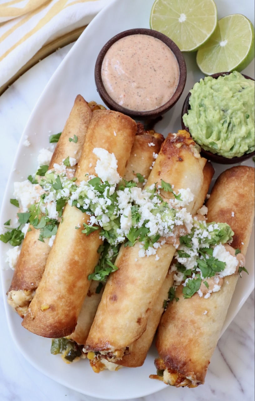 cooked taquitos stacked up on a plate topped with guacamole, cheese and cilantro