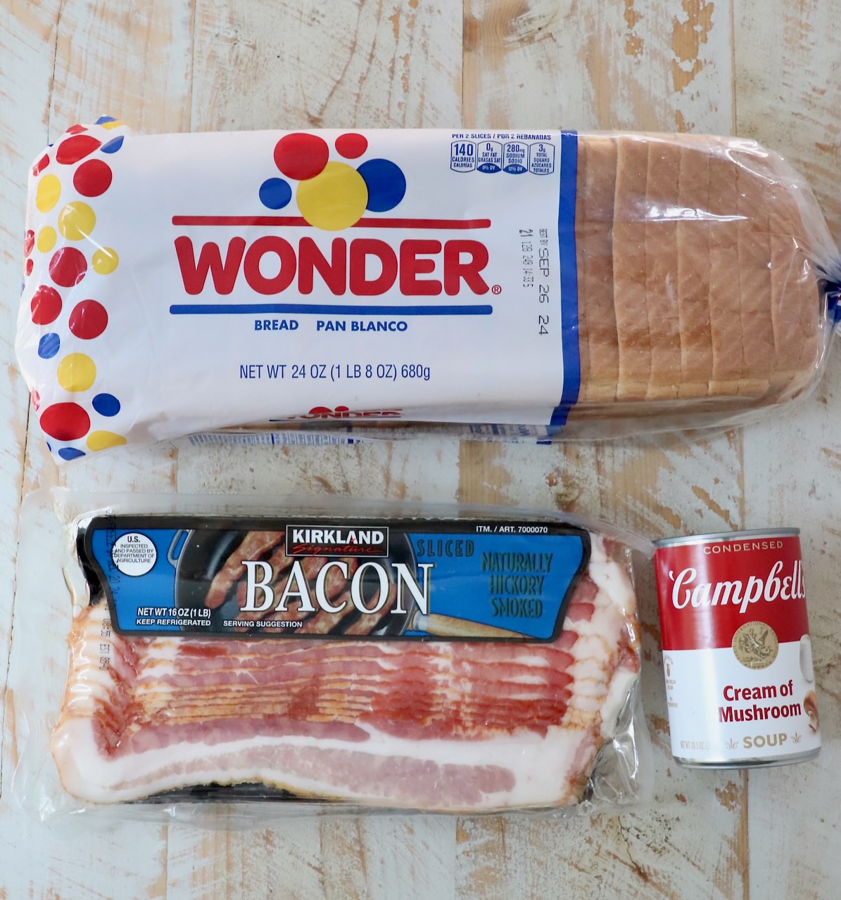 a package of bacon, loaf of wonder bread and a can of cream of mushroom soup, on a white wood board