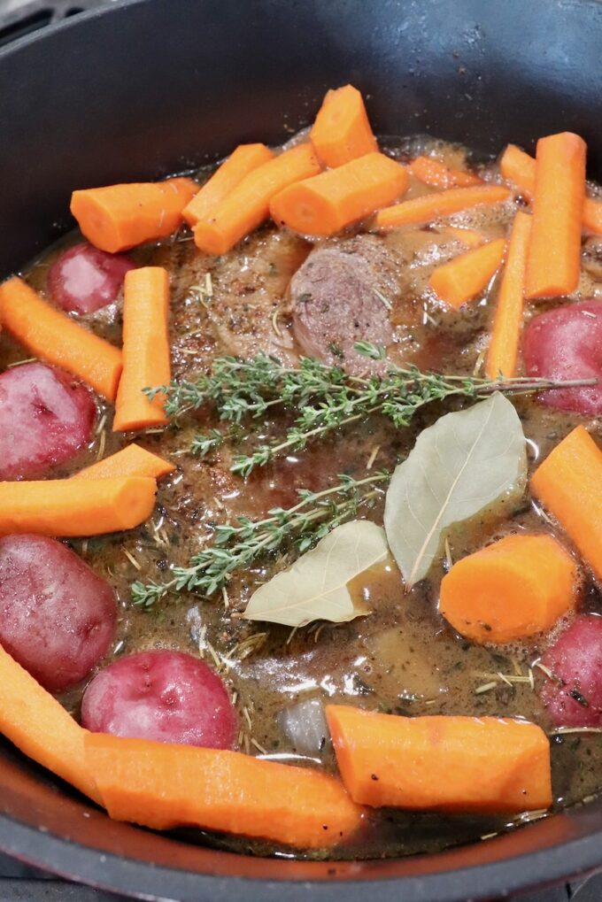 chuck roast in liquid in a dutch oven with carrots, potatoes, bay leaves and thyme sprigs