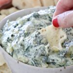 hand dipping a tortilla chip in a bowl of spinach artichoke dip