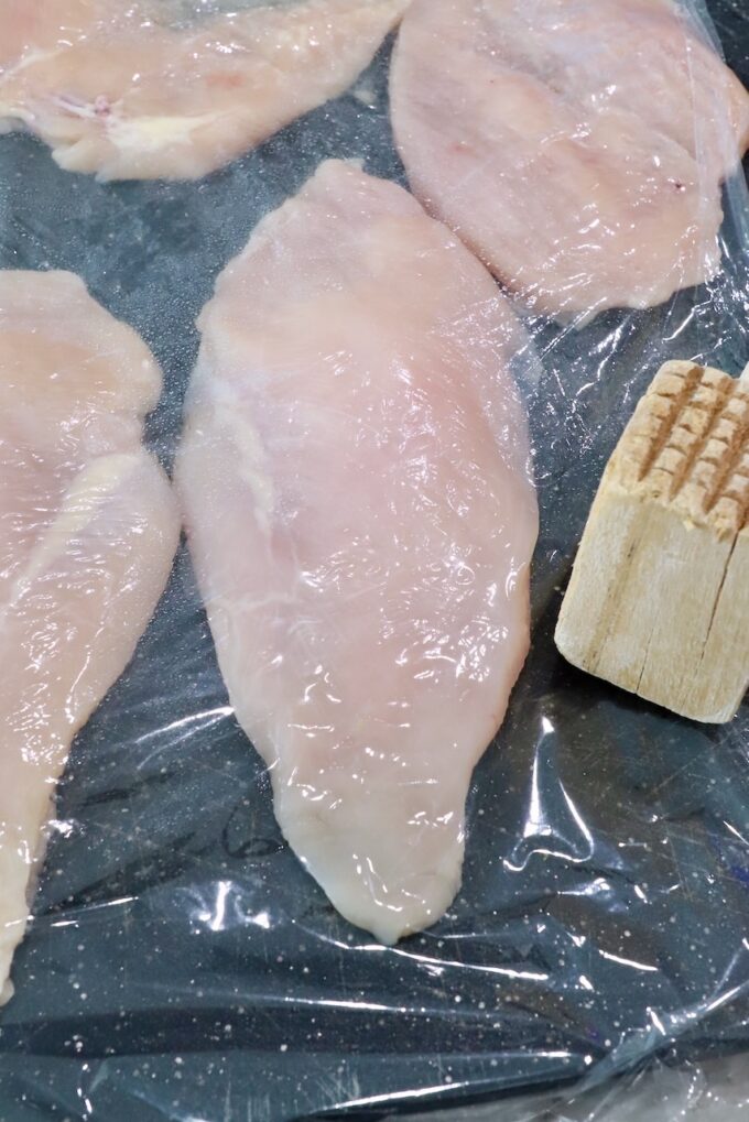 chicken breasts under plastic wrap on a cutting board