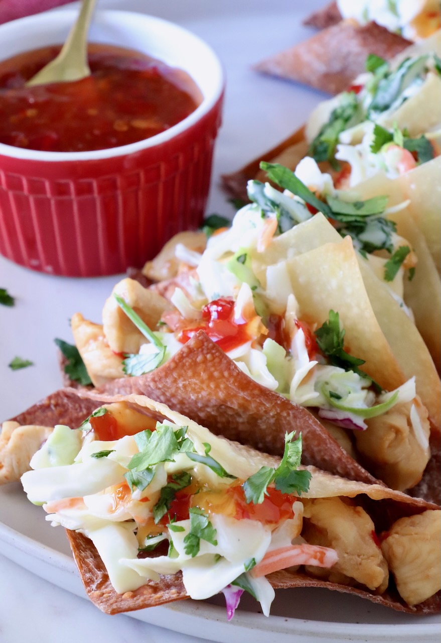 wonton tacos filled with chicken and coleslaw lined up on a plate next to a small bowl of sweet chili sauce