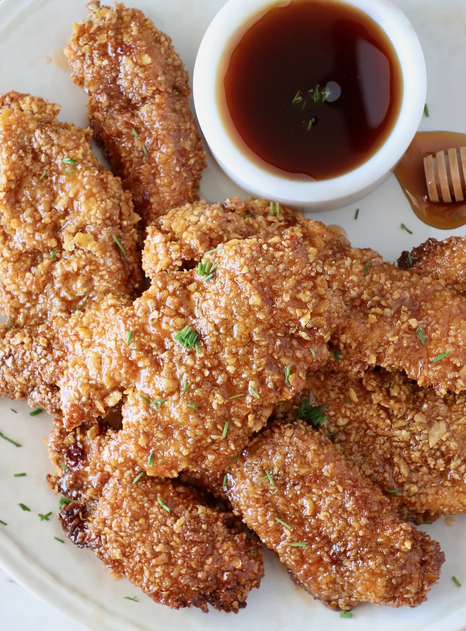 crispy chicken tenders piled up on a plate with a small bowl of hot honey sauce on the side