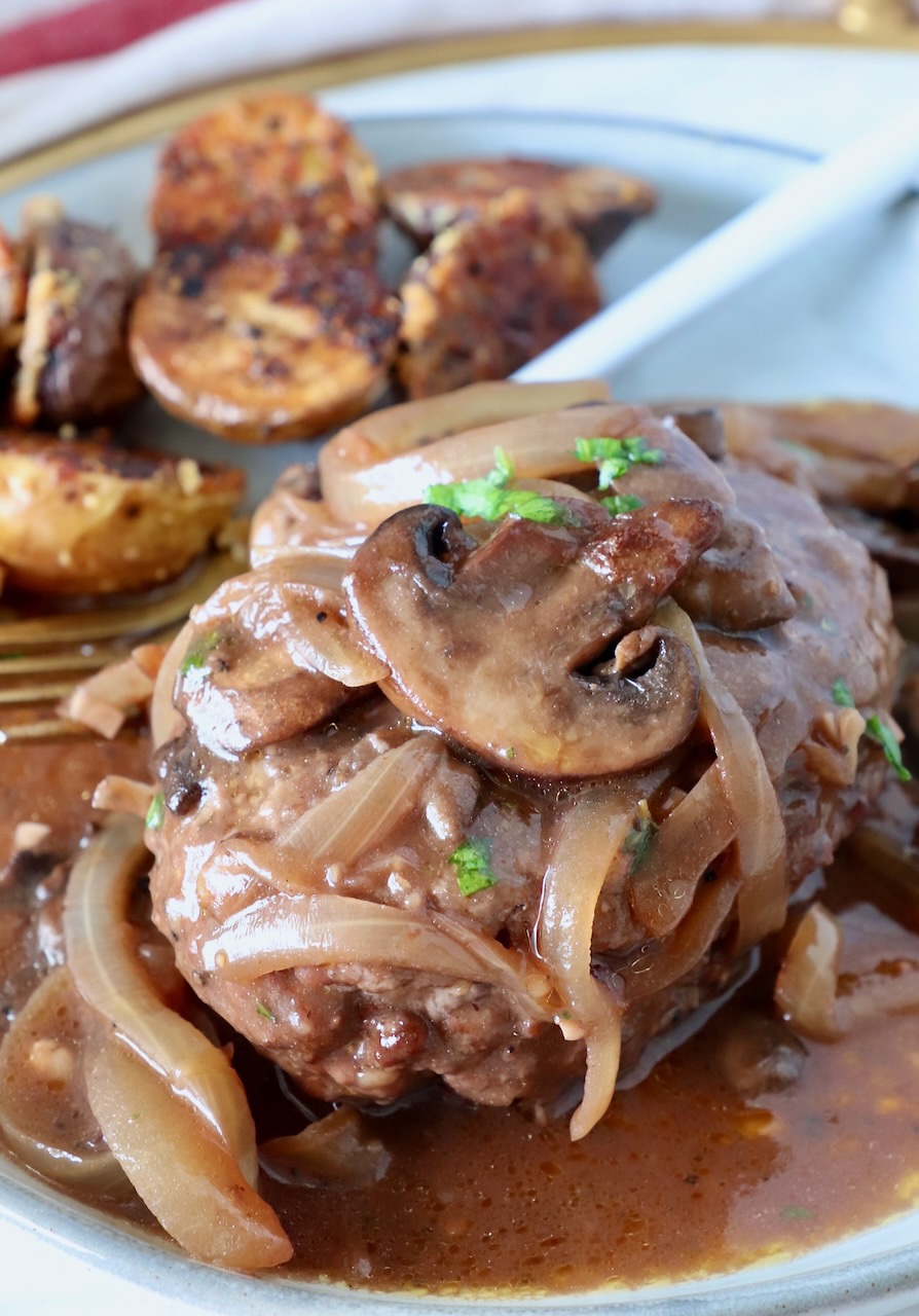 chopped steak on a plate covered with cooked mushrooms and onions, and red wine gravy, with roasted potatoes and a fork on the side of the plate