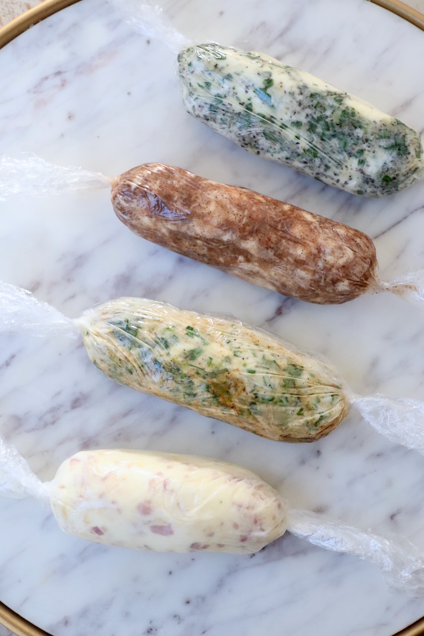 different types of compound butter wrapped up in plastic wrap on a marble serving tray