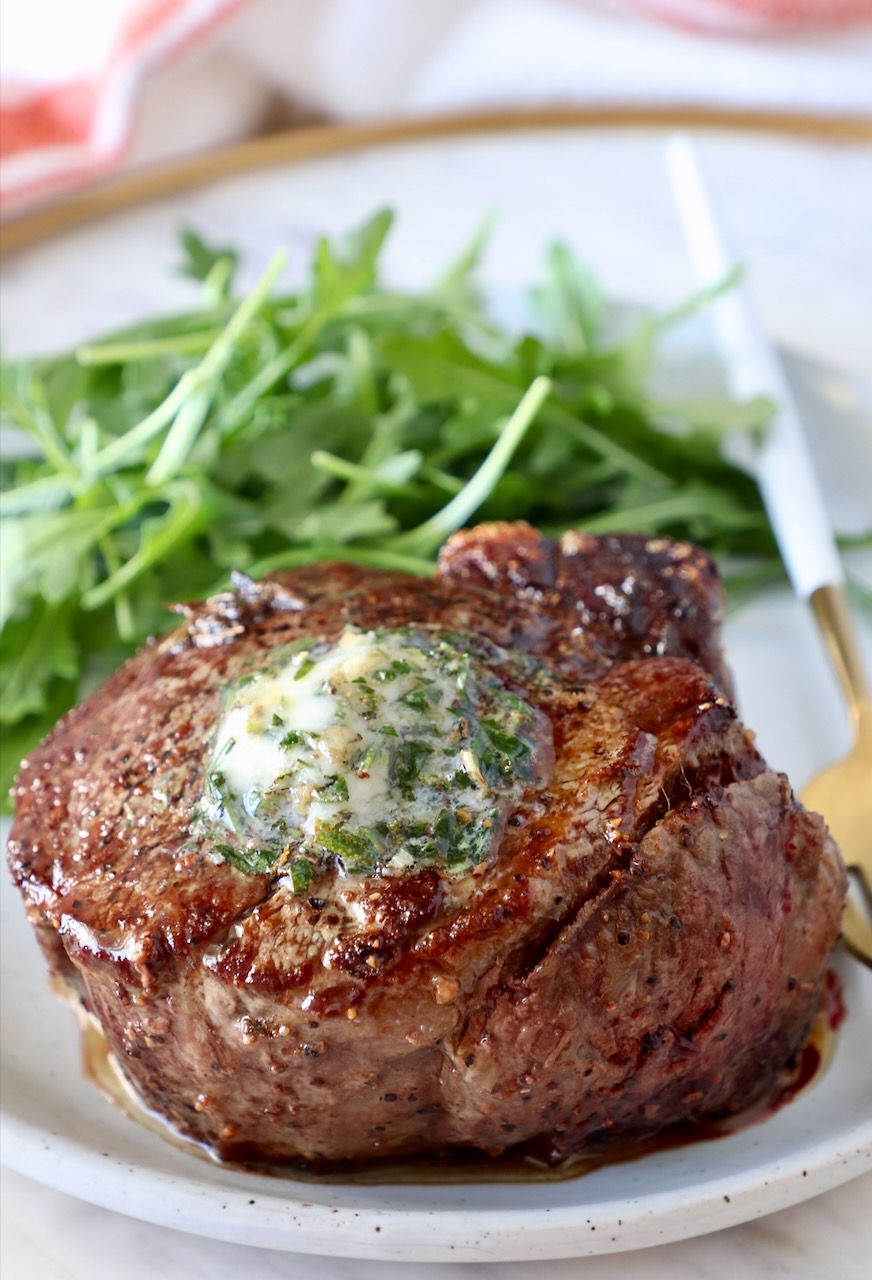 cooked steak on a plate topped with herb butter with arugula on the side of the plate