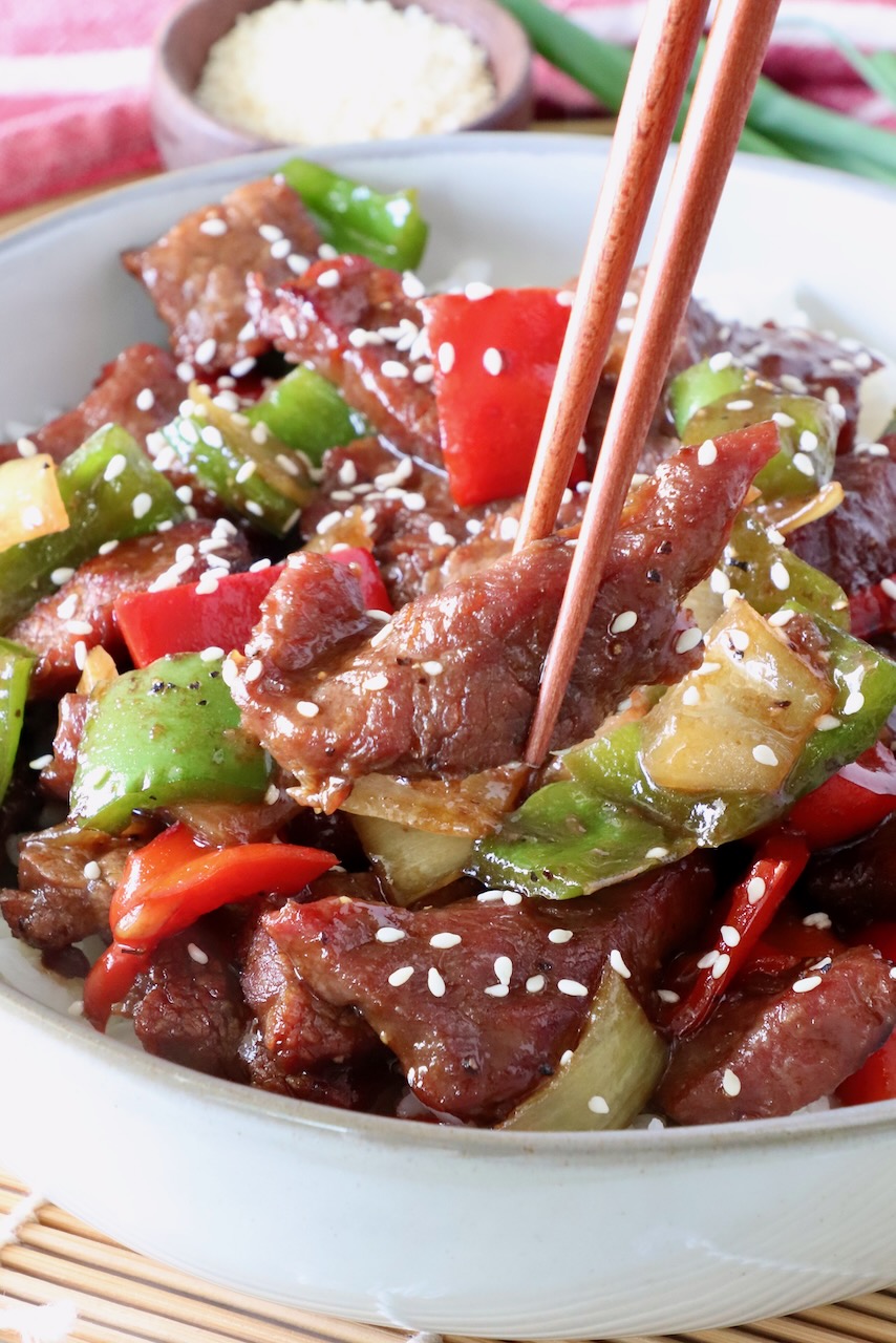 chopsticks holding a piece of steak over a bowl of cooked steak and peppers