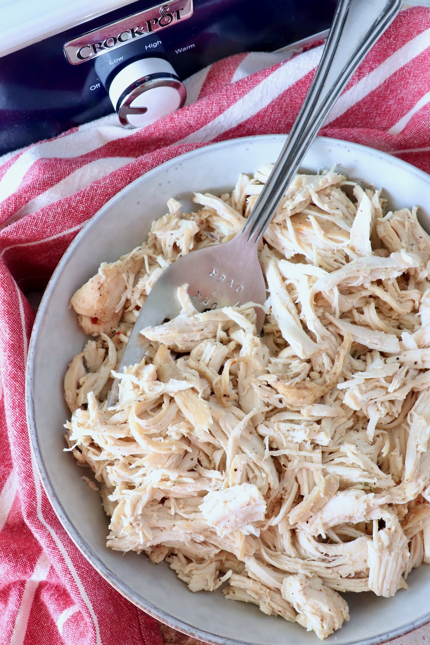 shredded chicken in a bowl with a large serving fork, sitting next to a red striped towel and a blue Crock Pot