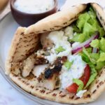 pita bread filled with chicken gyros meat, tzatziki sauce, shredded lettuce, sliced onions and tomatoes, on a plate