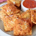 toasted raviolis stacked up on a plate with a small bowl of marinara sauce on the plate