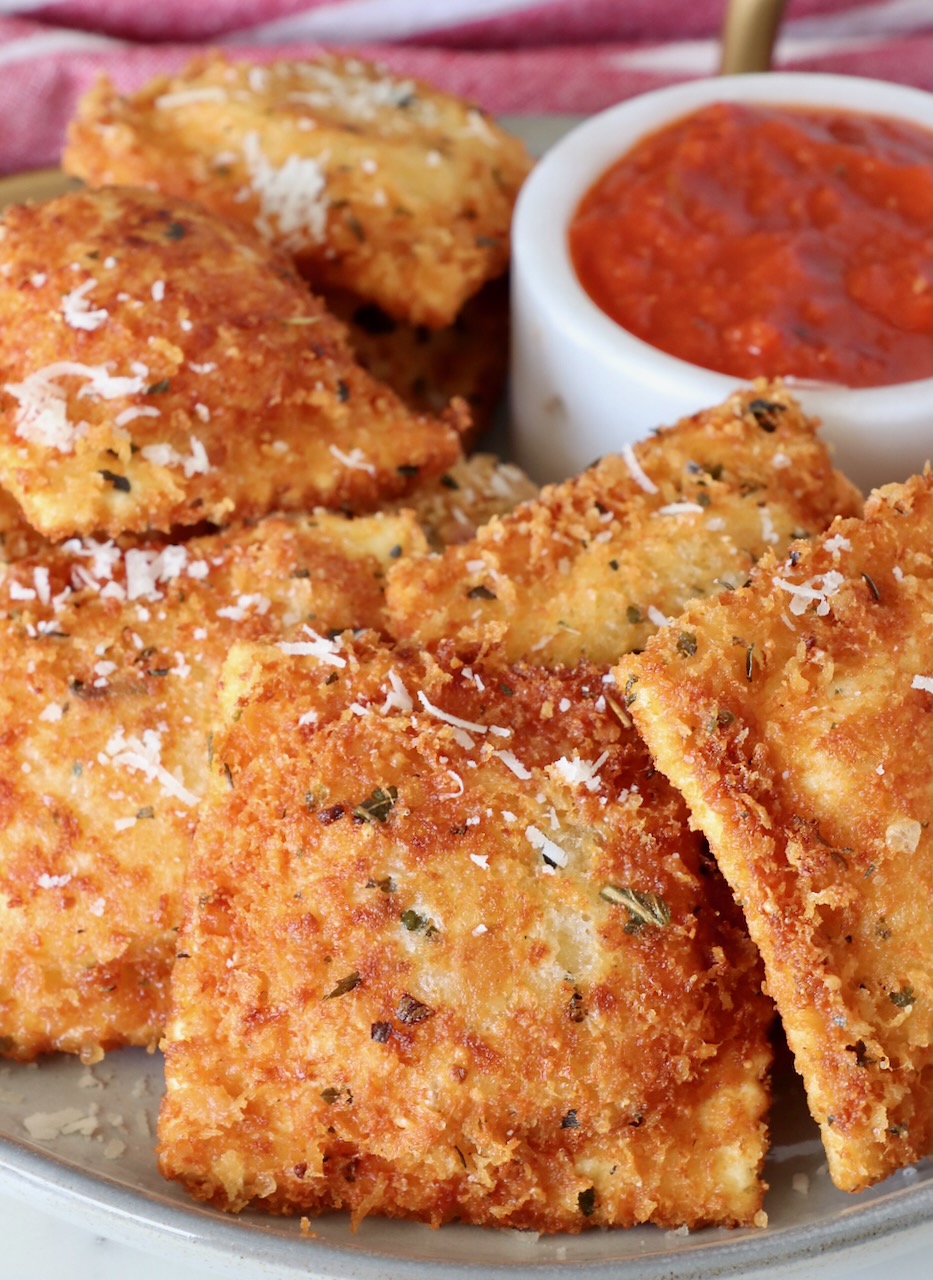 toasted raviolis stacked up on a plate with a bowl of marinara sauce behind them