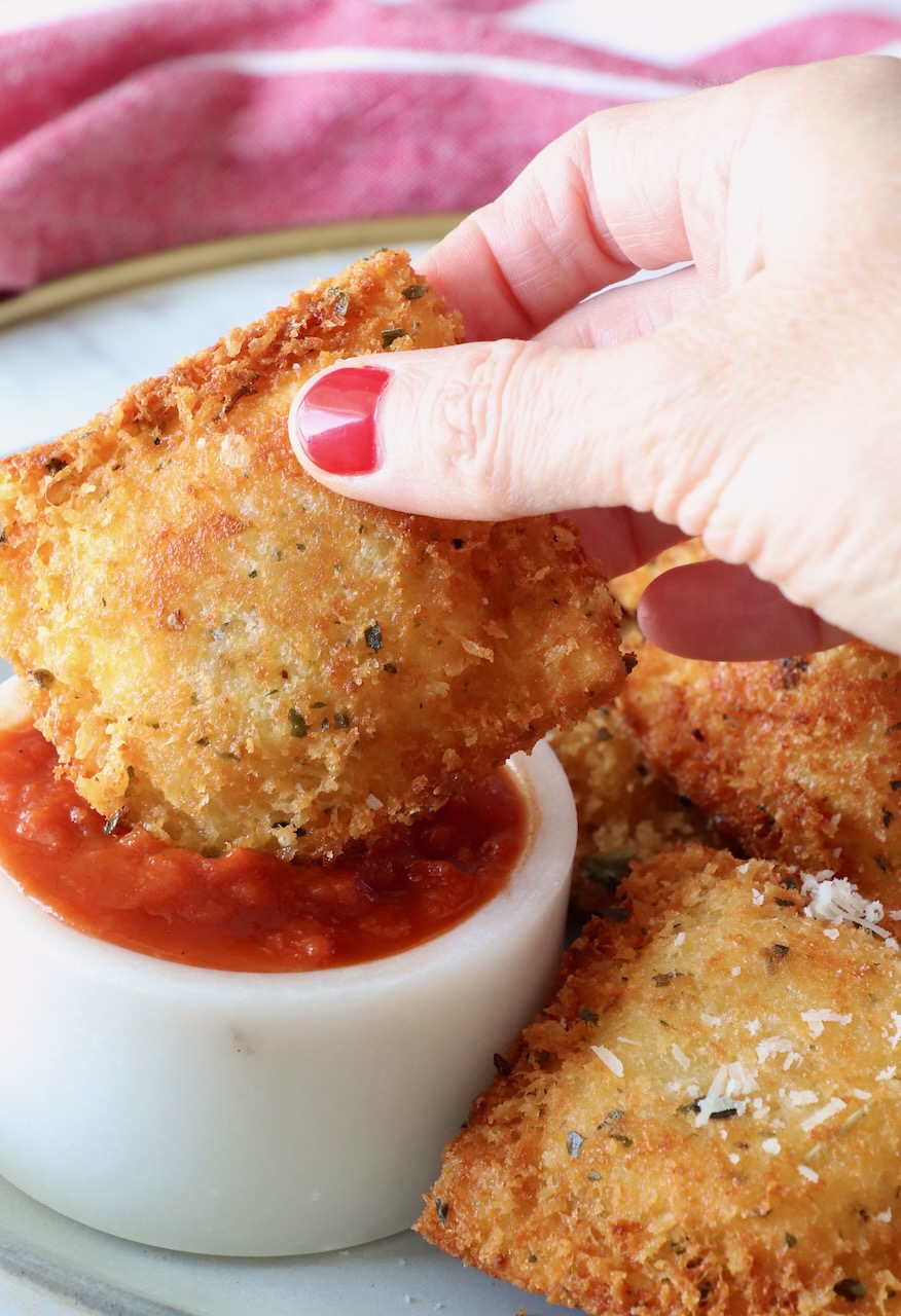 hand holding a toasted ravioli dipped into a small bowl of marinara sauce