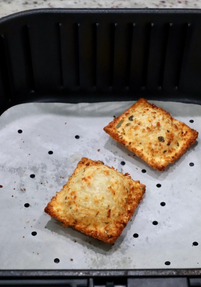 two toasted raviolis in an air fryer basket with a parchment paper liner