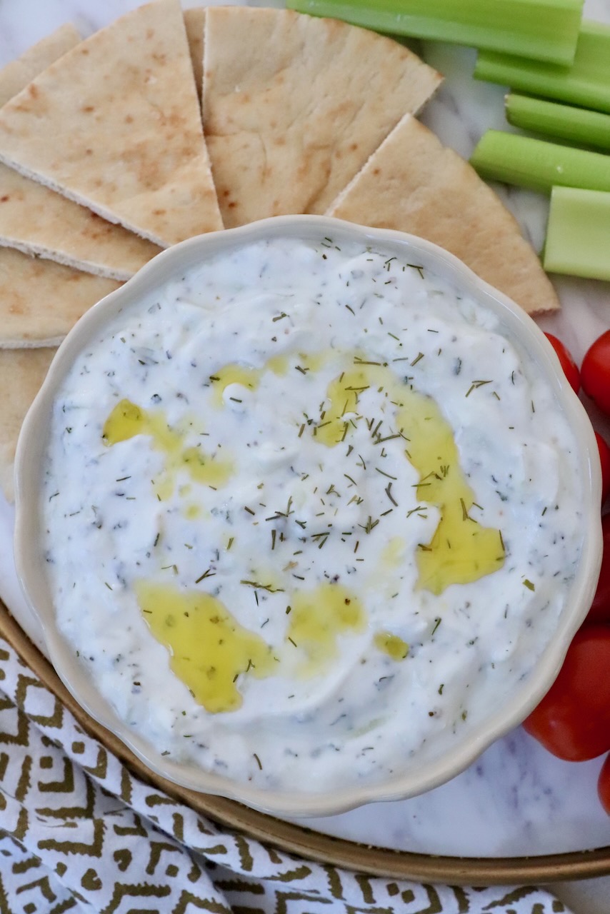 tzatziki dip in a bowl with olive oil drizzle on top, with sliced pita bread and vegetables on the side of the bowl